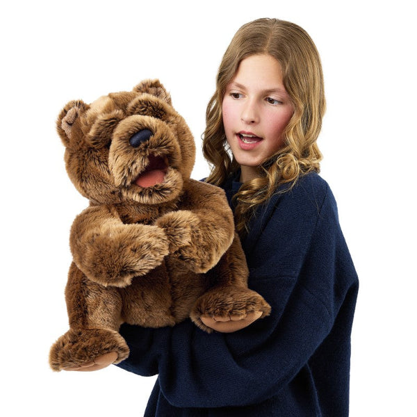A young girl with blonde hair wearing a navy blue sweater puppeteering a large, realistic brown plush bear puppet with a stitched open mouth and black nose.
