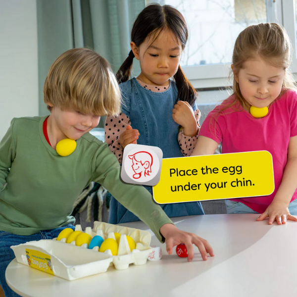 Three children playing with colorful eggs on a table, with a sign instructing them to place an egg under their chin.