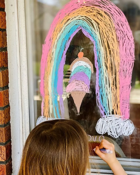 Child drawing a colorful rainbow on a window with crayons.